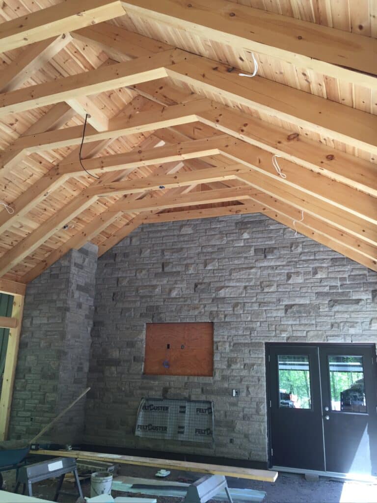 A room with a high, wooden vaulted ceiling features a stone accent wall and dark double doors. Construction materials are visible on the floor, and a covered fireplace is set into the stone wall. Natural light streams in through the windows.