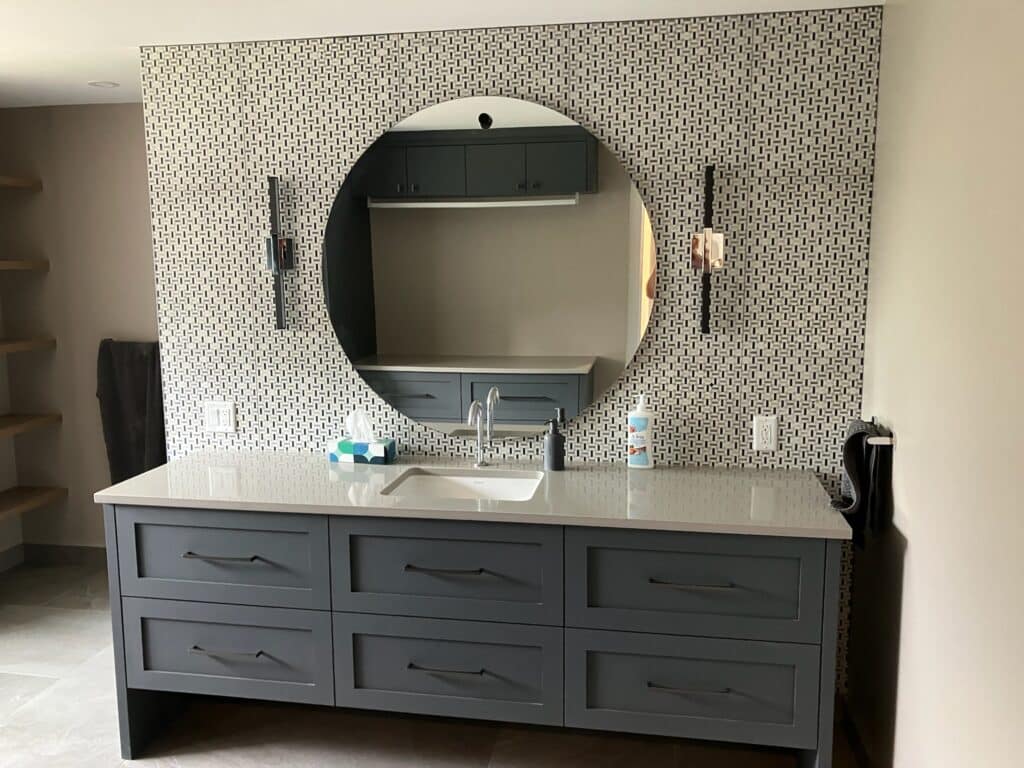 A modern bathroom vanity with a large round mirror on a patterned backsplash. The vanity has a white countertop, a sink, and multiple drawers with sleek handles. Two wall sconces are mounted beside the mirror.