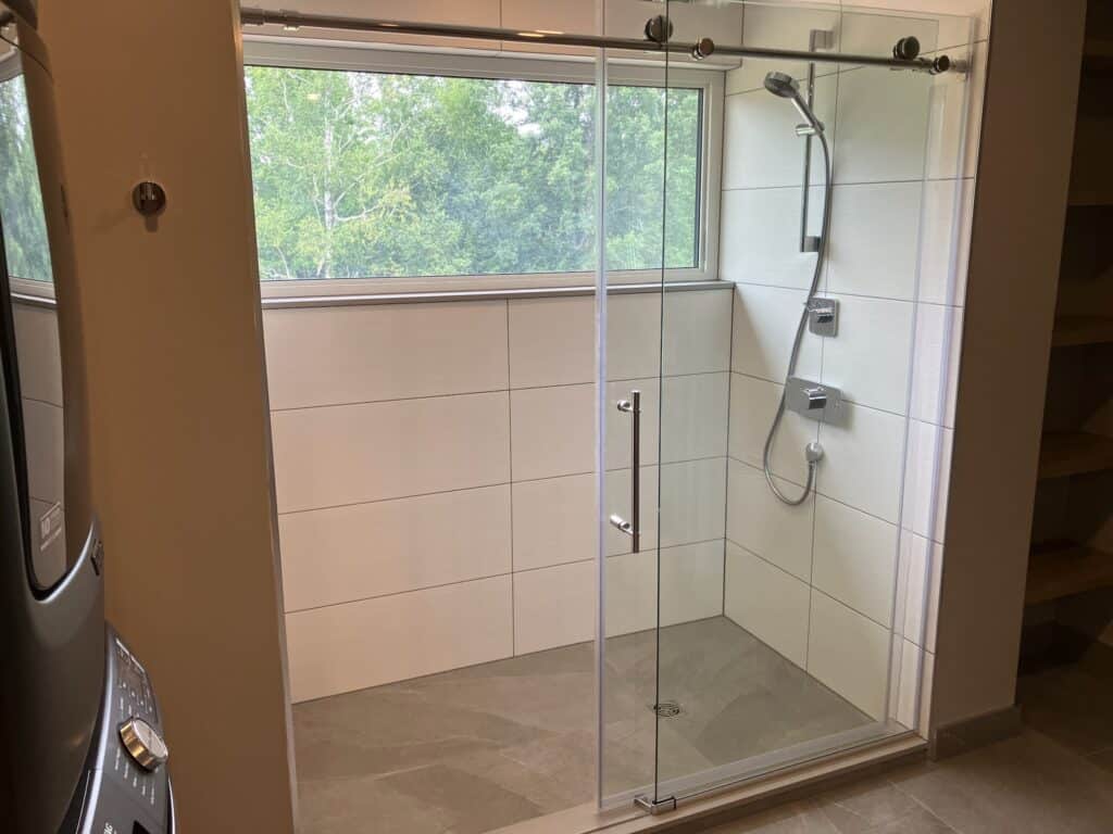 A modern bathroom with a glass-enclosed shower. The shower features a handheld showerhead and built-in shelves. Light-colored tiles line the walls, and a window above the shower provides natural light and a view of green foliage outside.