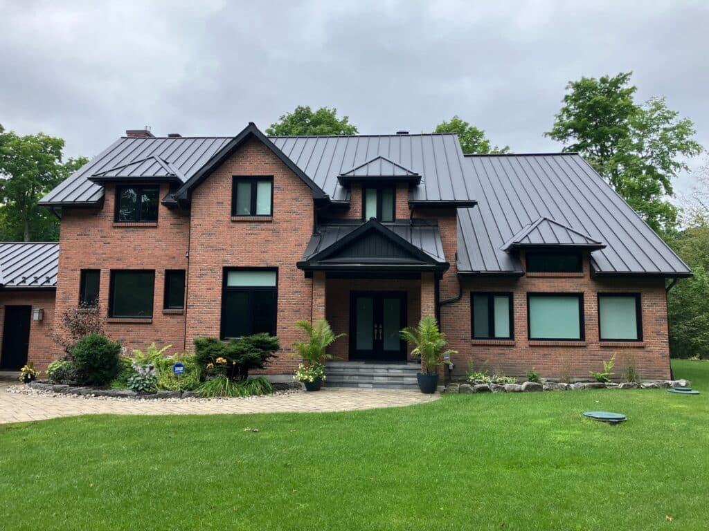 A two-story brick house with a dark metal roof, surrounded by a well-maintained lawn and trees. The entrance features potted plants on either side, and the sky is overcast.