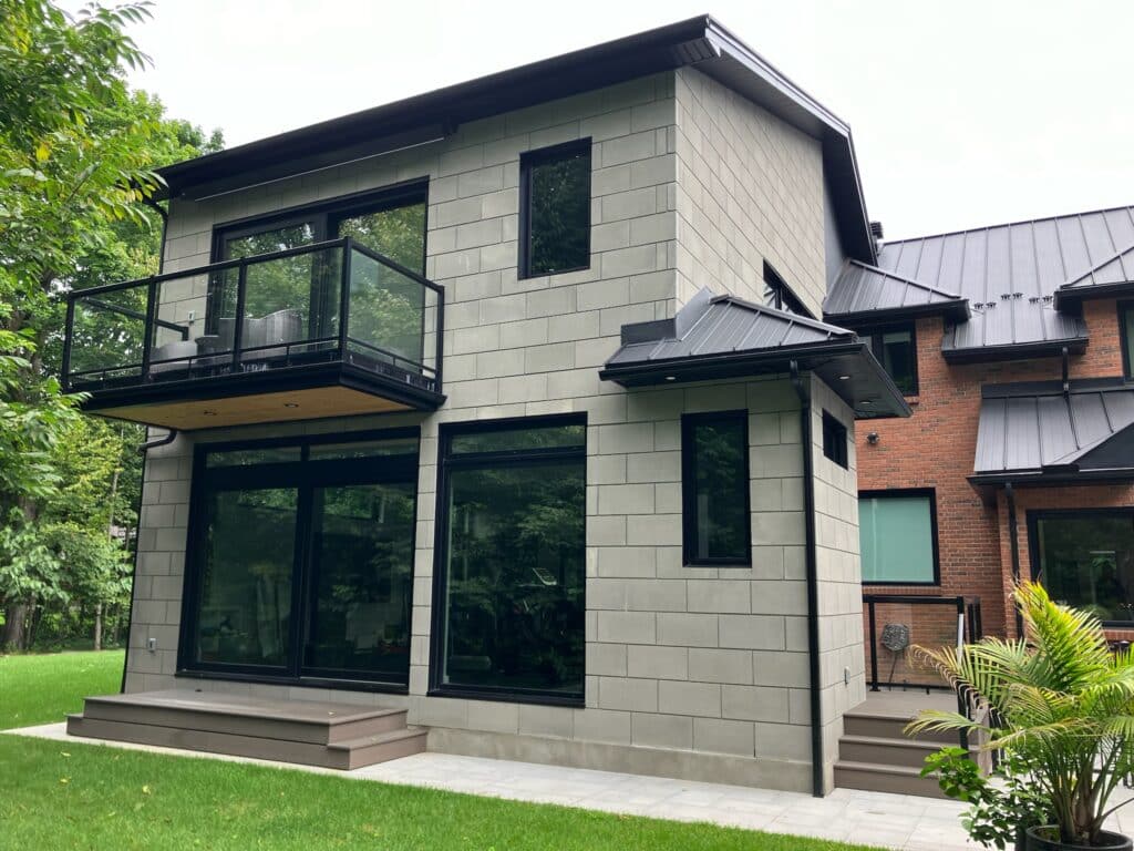 Modern two-story house with large windows and a second-floor balcony. The exterior features a mix of gray and brick walls, surrounded by a grassy yard and trees. Steps lead up to the glass doors, with a potted plant on the right.