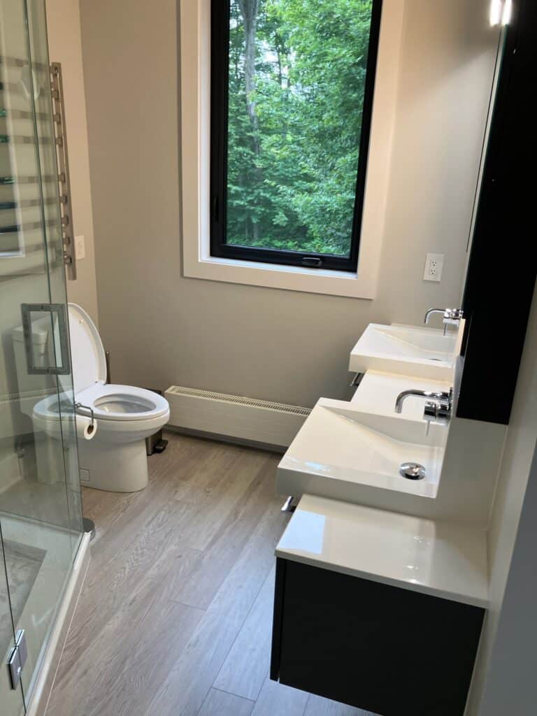 A modern bathroom with a glass shower enclosure, two white sinks on a floating vanity, and a toilet next to a large window overlooking a green forest. The floor is light wood, and the walls are painted in a soft neutral tone.