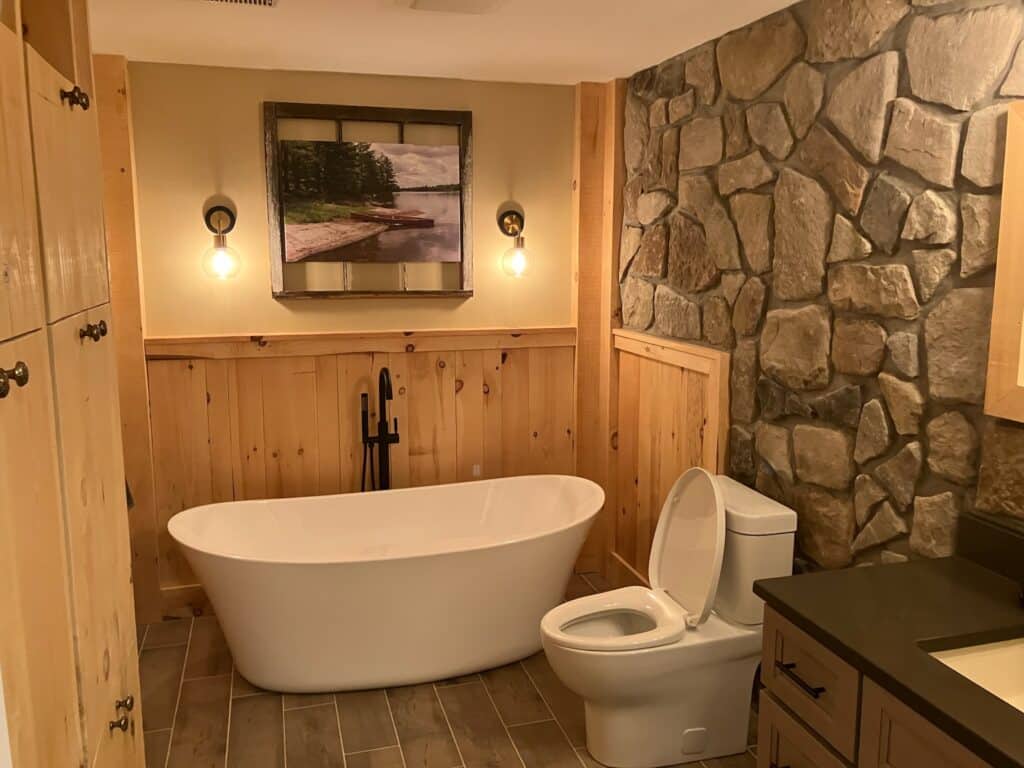 A rustic bathroom features a freestanding white tub, modern toilet, and stone-accented wall. Wooden paneling adds warmth, while two wall lights illuminate a nature-themed picture. A dark sink countertop complements the natural aesthetic.