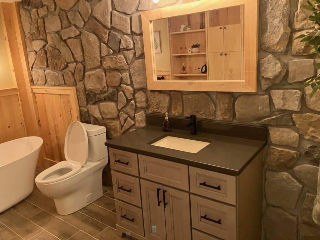 A cozy bathroom with a rustic stone wall and wooden accents. It features a white toilet, a freestanding bathtub, a vanity with a black countertop, a square mirror with a wooden frame, and a set of shelves. Warm lighting creates an inviting atmosphere.