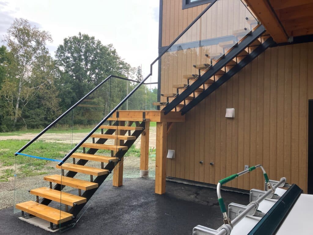 A set of modern outdoor wooden stairs with glass railings leads to the second floor of a building. The building has vertical wood siding. The background features trees and a grassy area under an overcast sky.