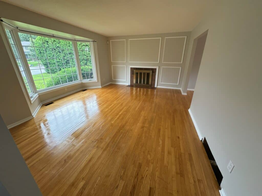 A spacious, empty living room with polished wooden floors and large windows offering a view of greenery outside. The room features a white-framed fireplace set into a wall with decorative molding. Natural light illuminates the space.