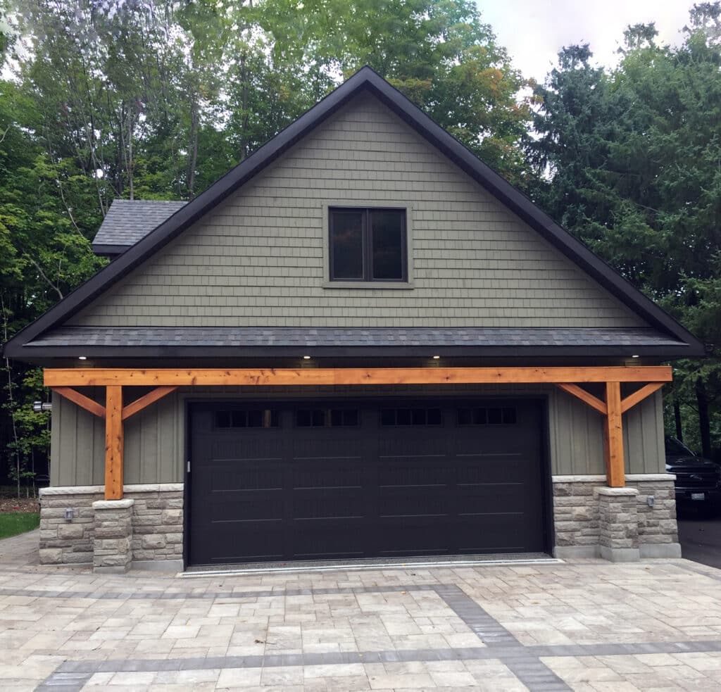 A modern garage with a pitched roof and dark brown double doors is set against a backdrop of trees. The structure features stone and wood accents, with a paved driveway in the foreground.