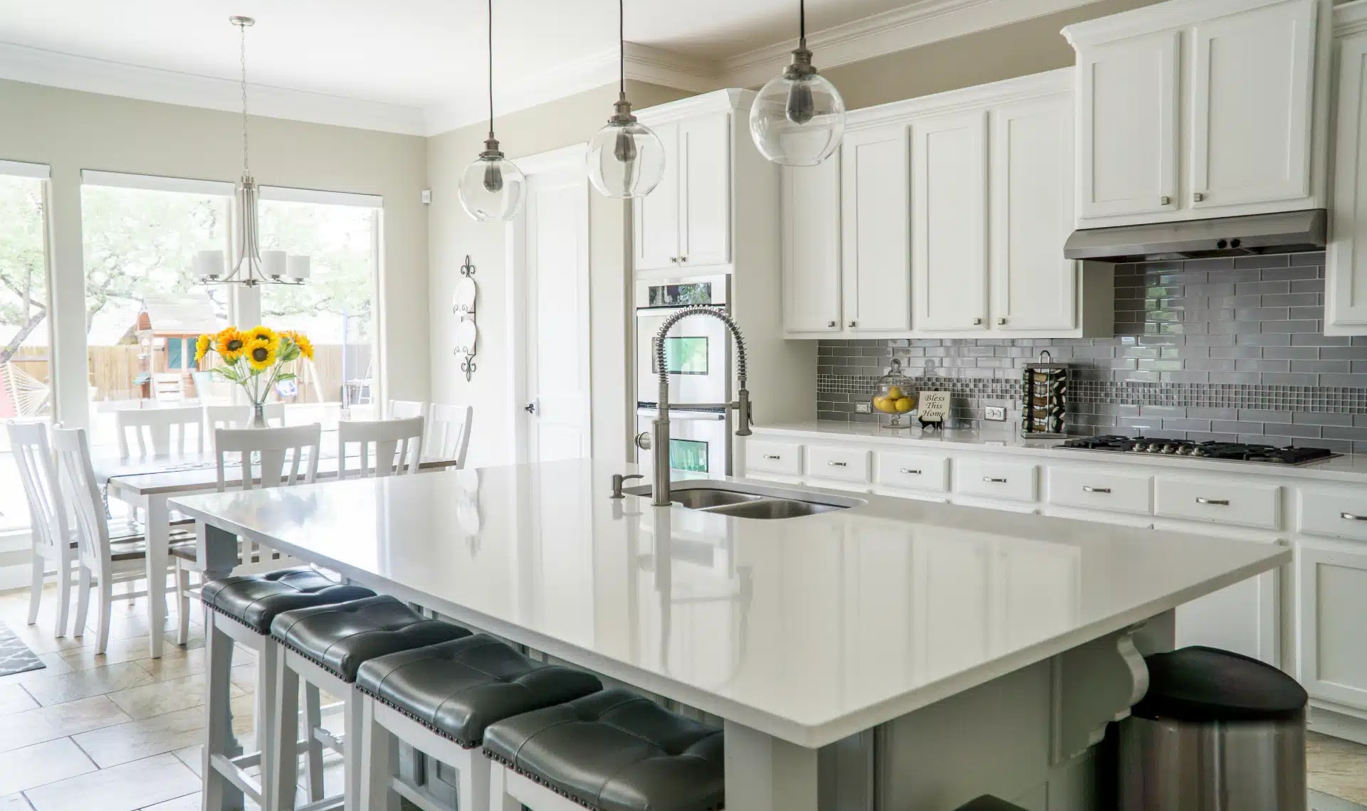 A modern kitchen with white cabinets, a large central island, and black cushioned stools. Stainless steel appliances, a gray tile backsplash, and hanging pendant lights add style. A vase of sunflowers sits on the dining table by the window.