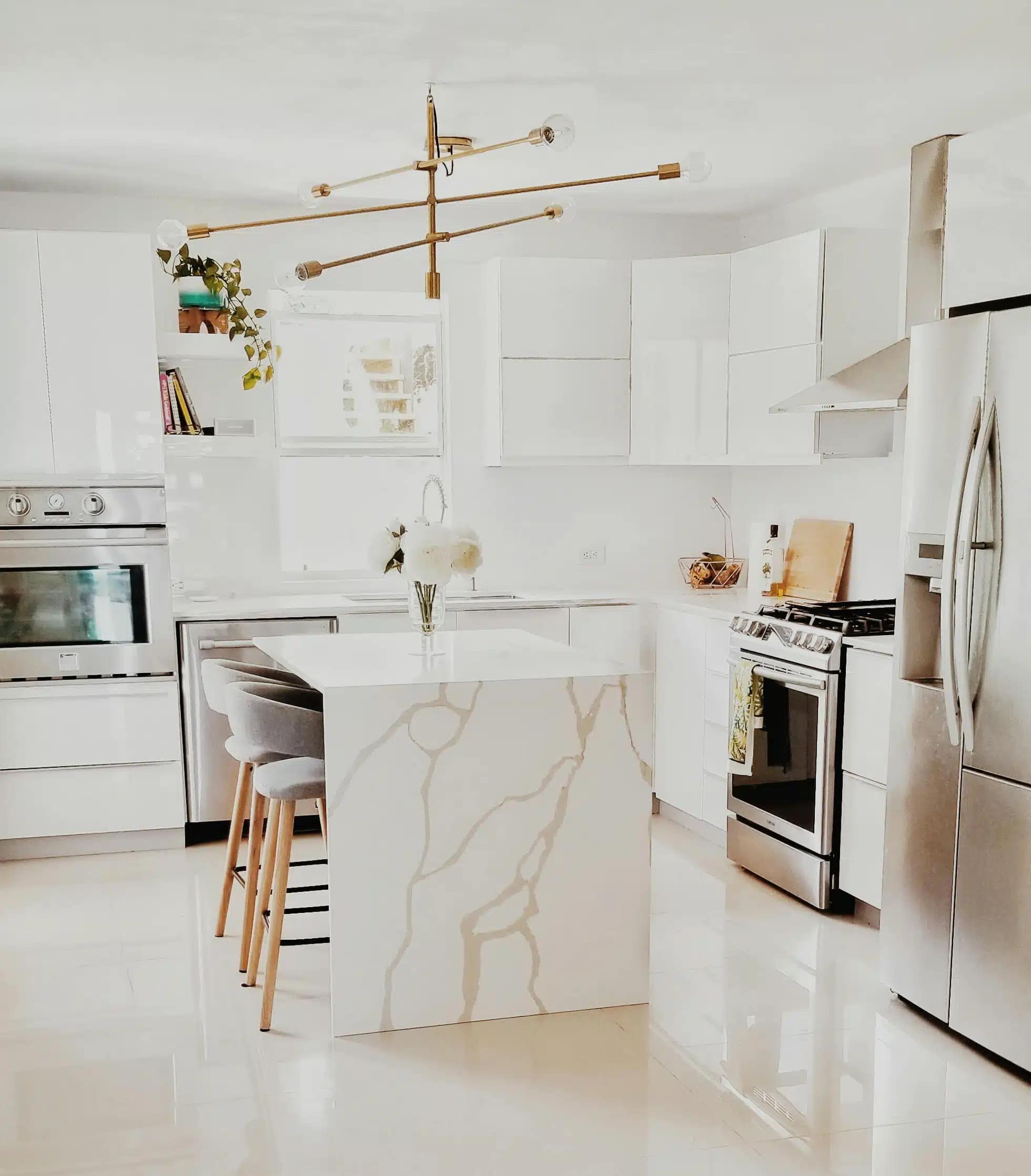 A modern kitchen with white cabinets and appliances. A marble island with gray stools is centered under a gold chandelier. The space is bright and minimalistic with flowers and plants for decoration.